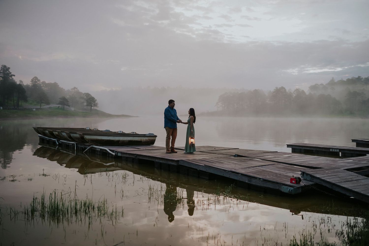 Silhouettes of people stand on a misty dock overlooking a tranquil lake at dawn.
