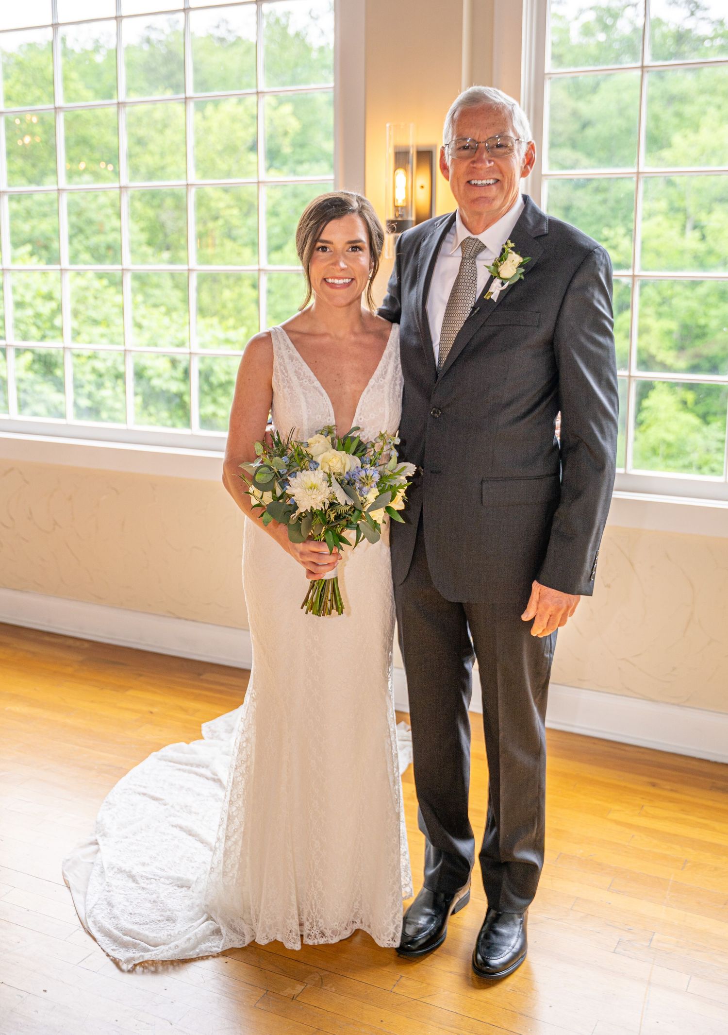 A bride in a white sheath dress and her companion in a grey suit pose by bright windows during a wedding celebration.