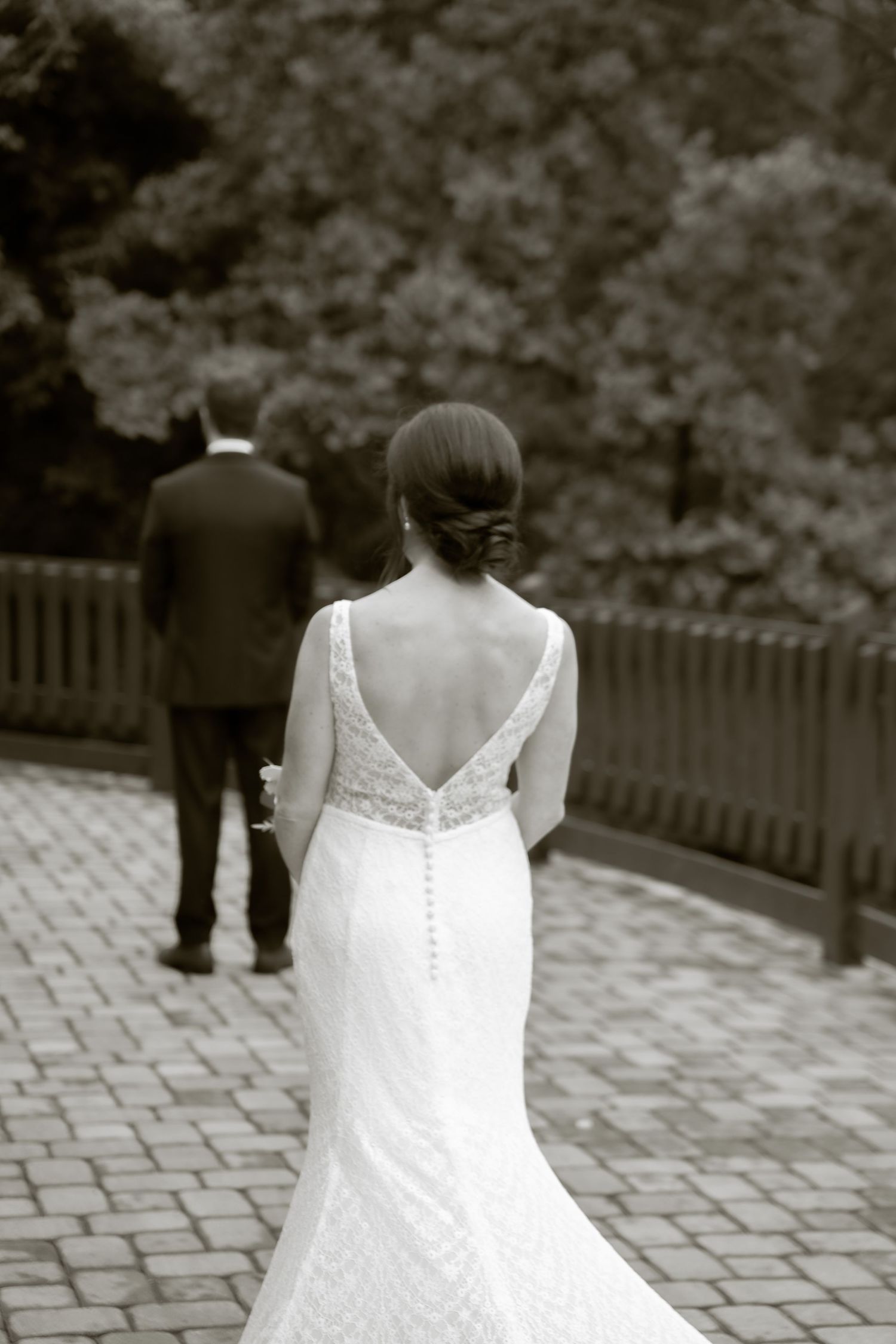 Black and white wedding photo showing the back view of a bride in an elegant button-back dress on cobblestone path.