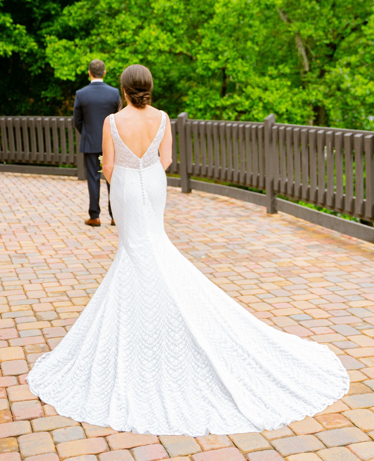 Elegant white mermaid wedding gown with intricate lace pattern and dramatic train on brick paved walkway with greenery backdrop.