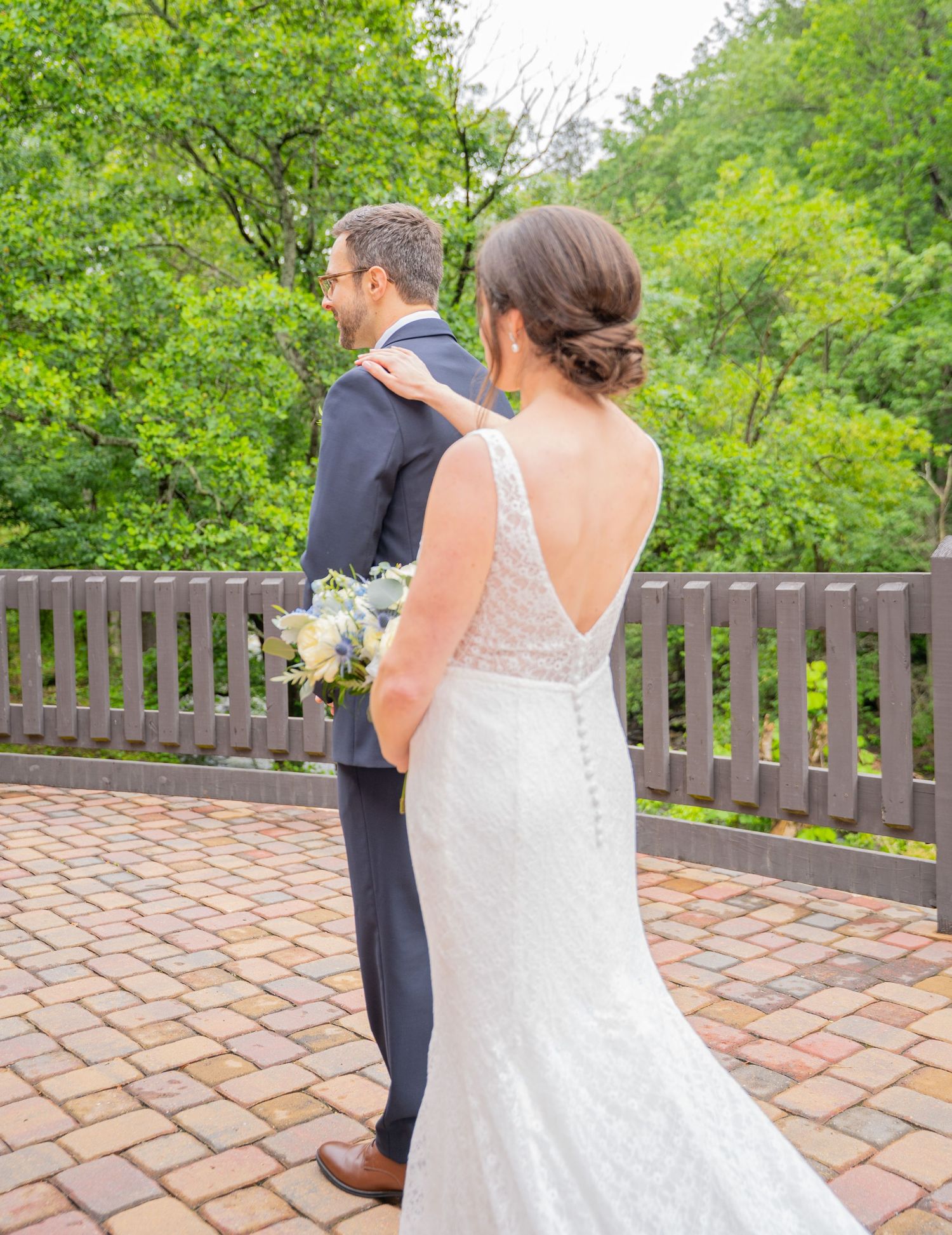 Bride in lace wedding dress and groom in navy suit share a first look moment on brick patio surrounded by greenery.