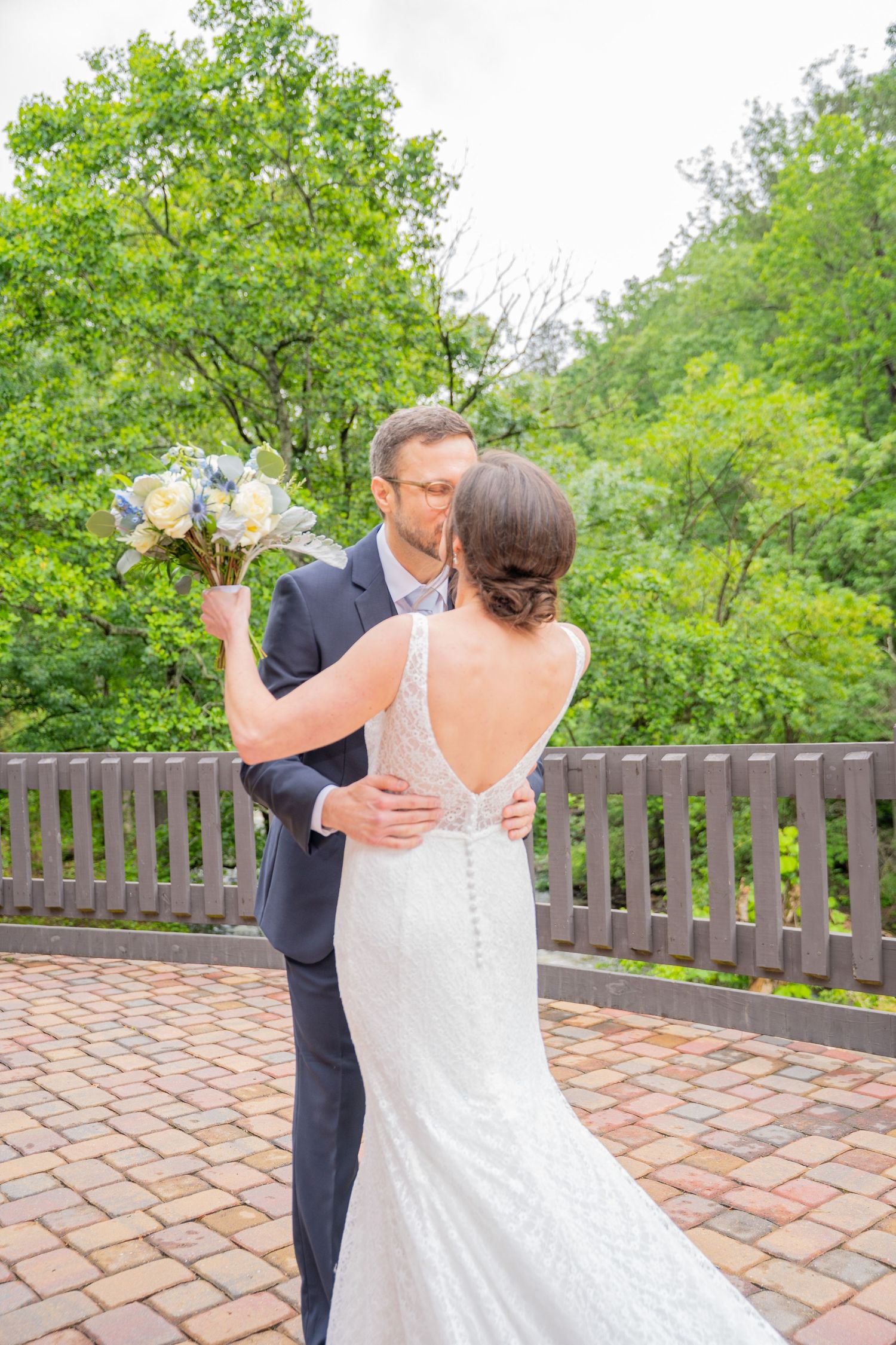 A couple shares an intimate moment outdoors on a brick patio with lush greenery and a wooden fence in the background.