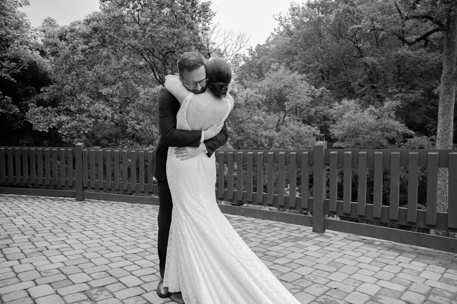 A black and white wedding photograph showing a couple embracing on a brick patio surrounded by trees and a wooden fence.