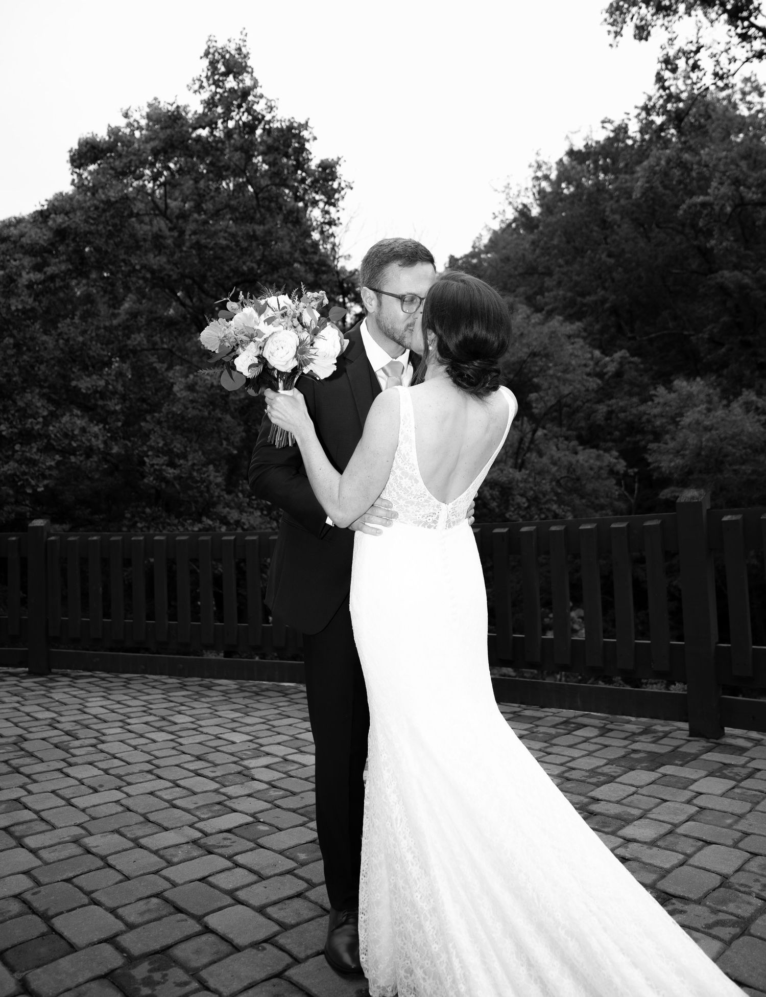 A tender wedding moment captured in black and white on a brick patio with lush trees in the background.