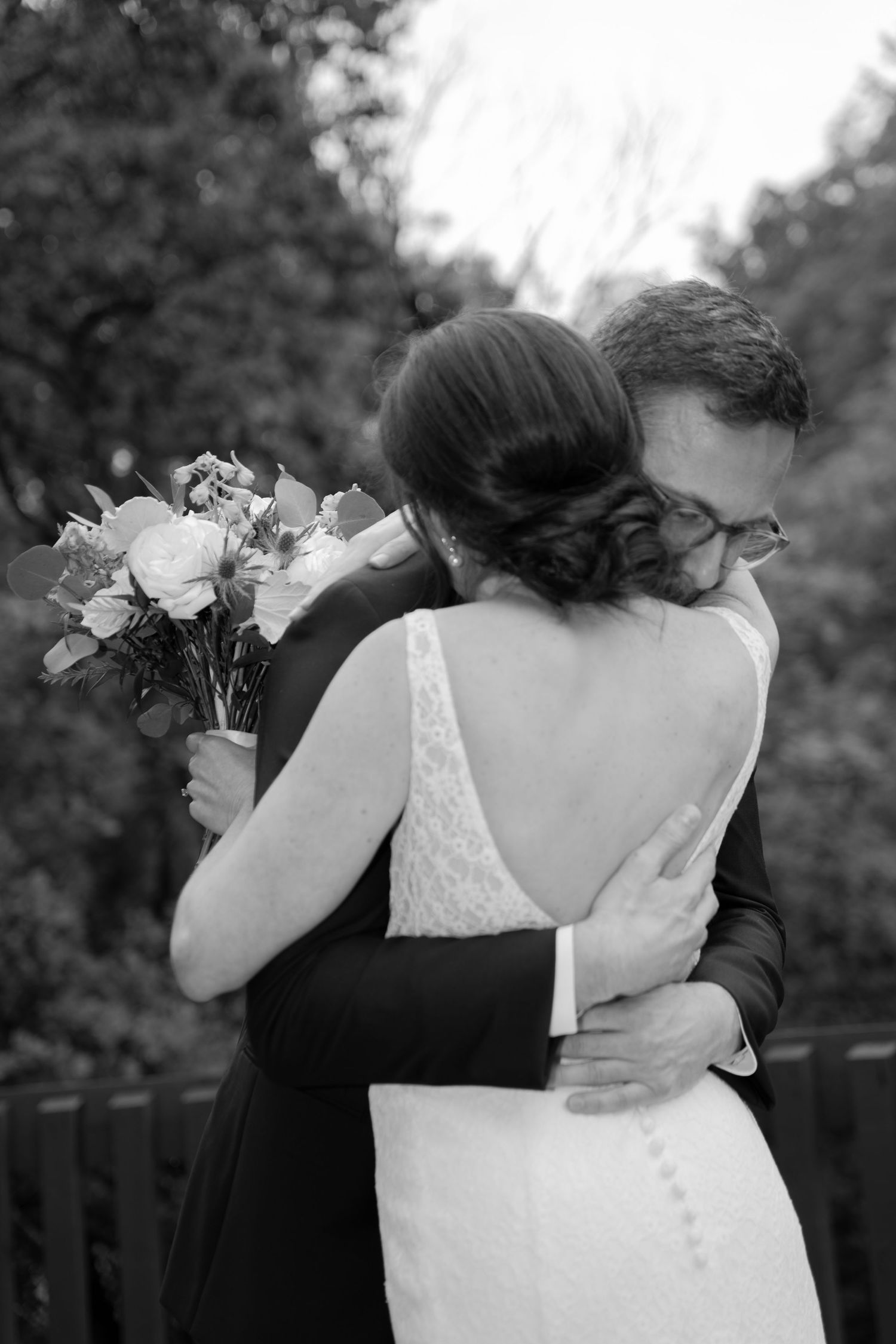 A romantic black and white photograph of a couple embracing during their wedding, with the bride holding a floral bouquet.