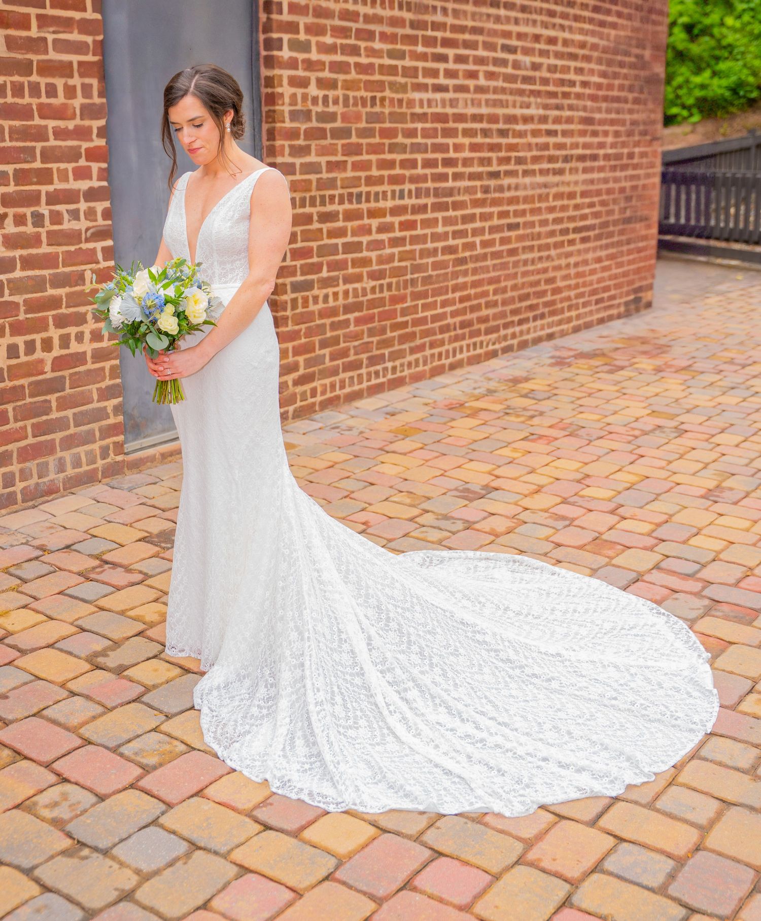 Elegant lace wedding gown with dramatic cathedral train displayed against rustic brick wall backdrop on cobblestone patio.