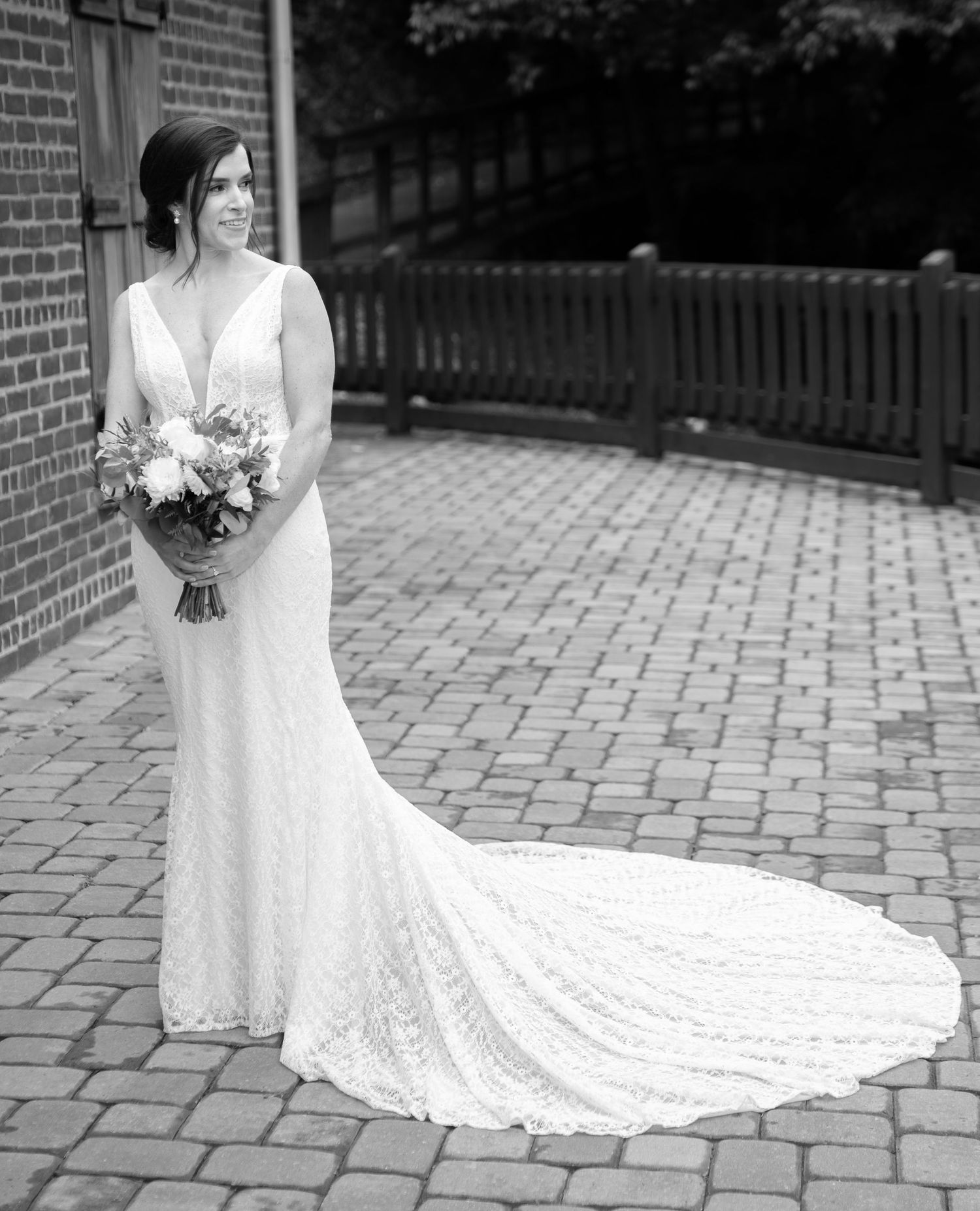 A bride in a long lace wedding dress with train holds a bouquet while standing on brick pavement outdoors.