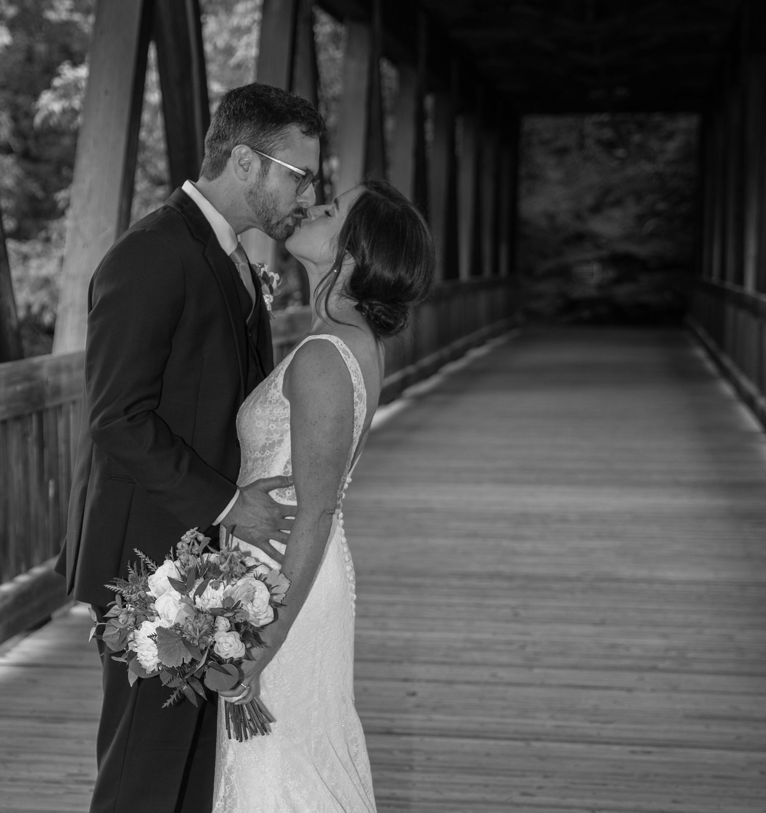 A romantic black and white wedding photo of a couple sharing an intimate moment on a covered wooden bridge.