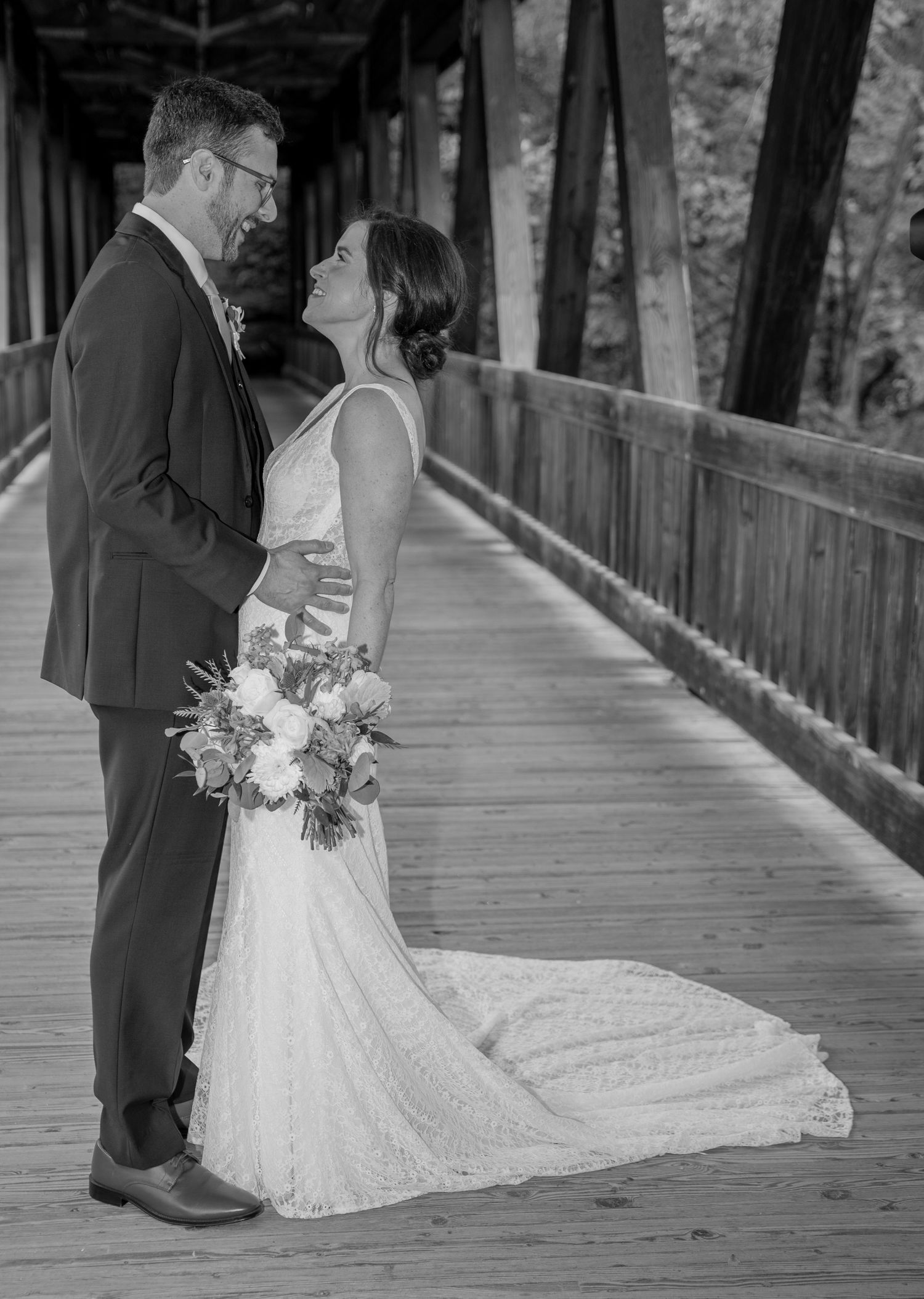 Black and white wedding photo of couple sharing intimate moment on wooden covered bridge holding floral bouquet.