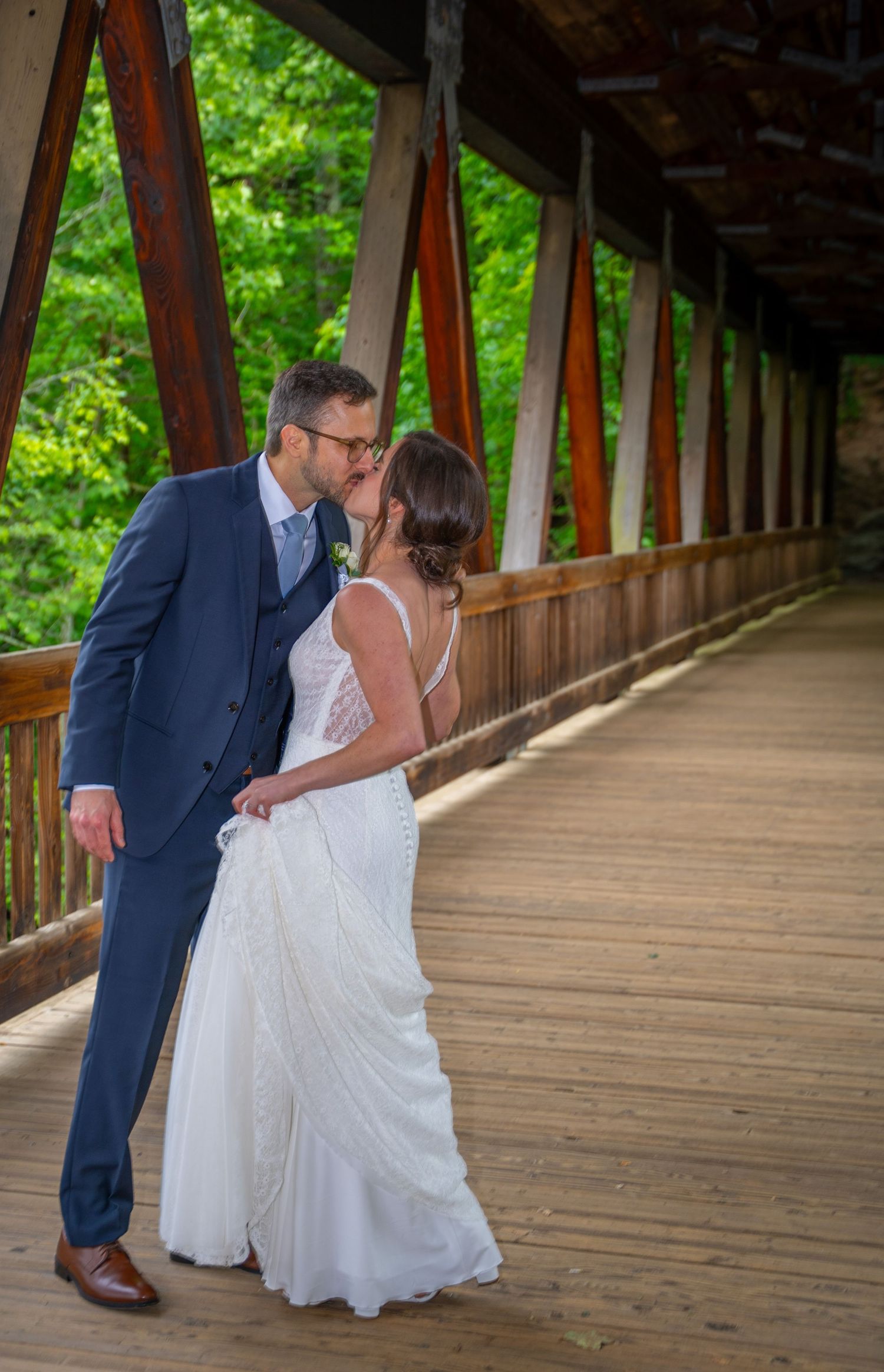 Romantic couple share a tender moment on a wooden covered bridge surrounded by lush green foliage.