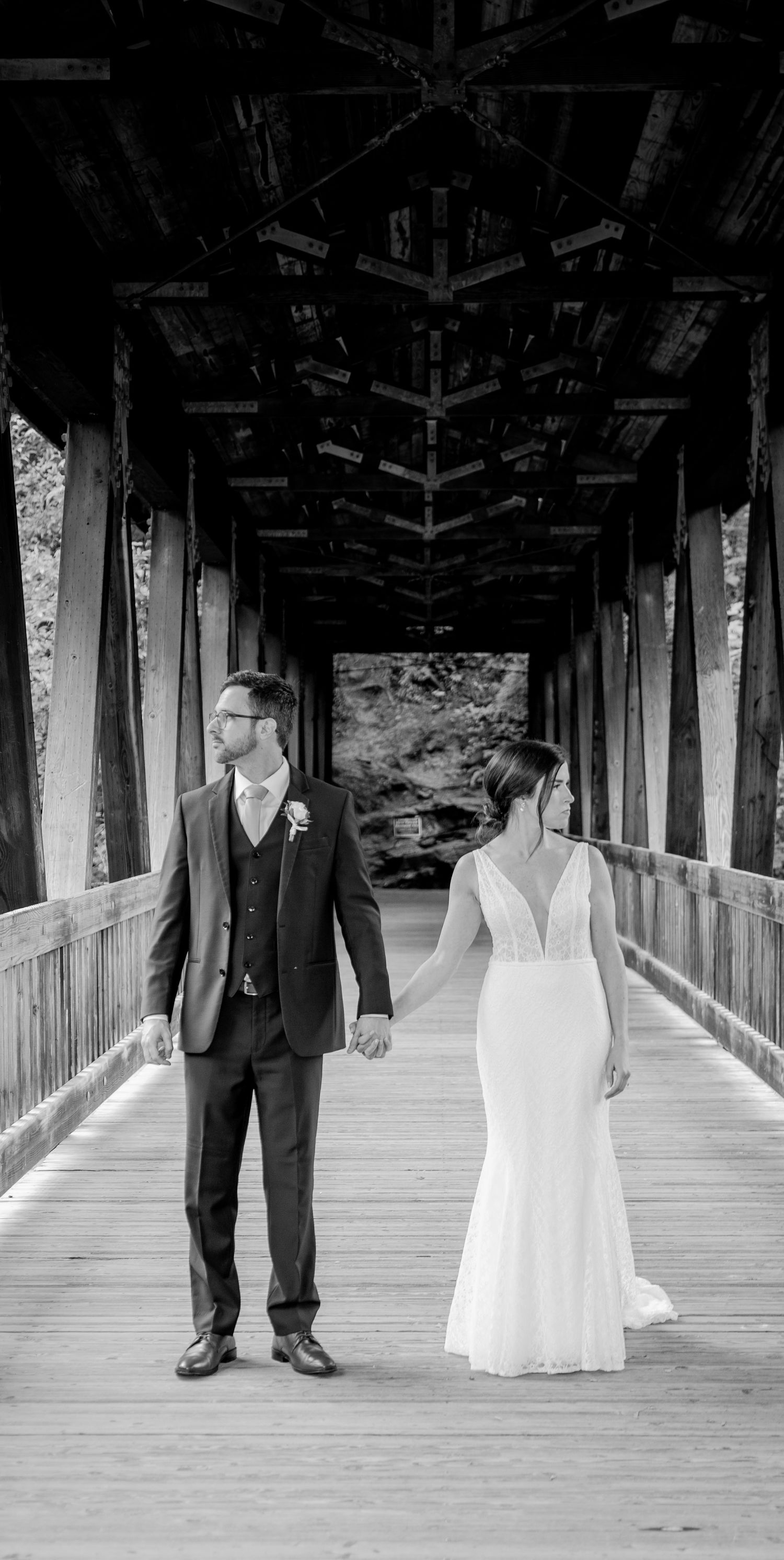 A couple in wedding attire stands back-to-back on a covered wooden bridge in black and white photography.
