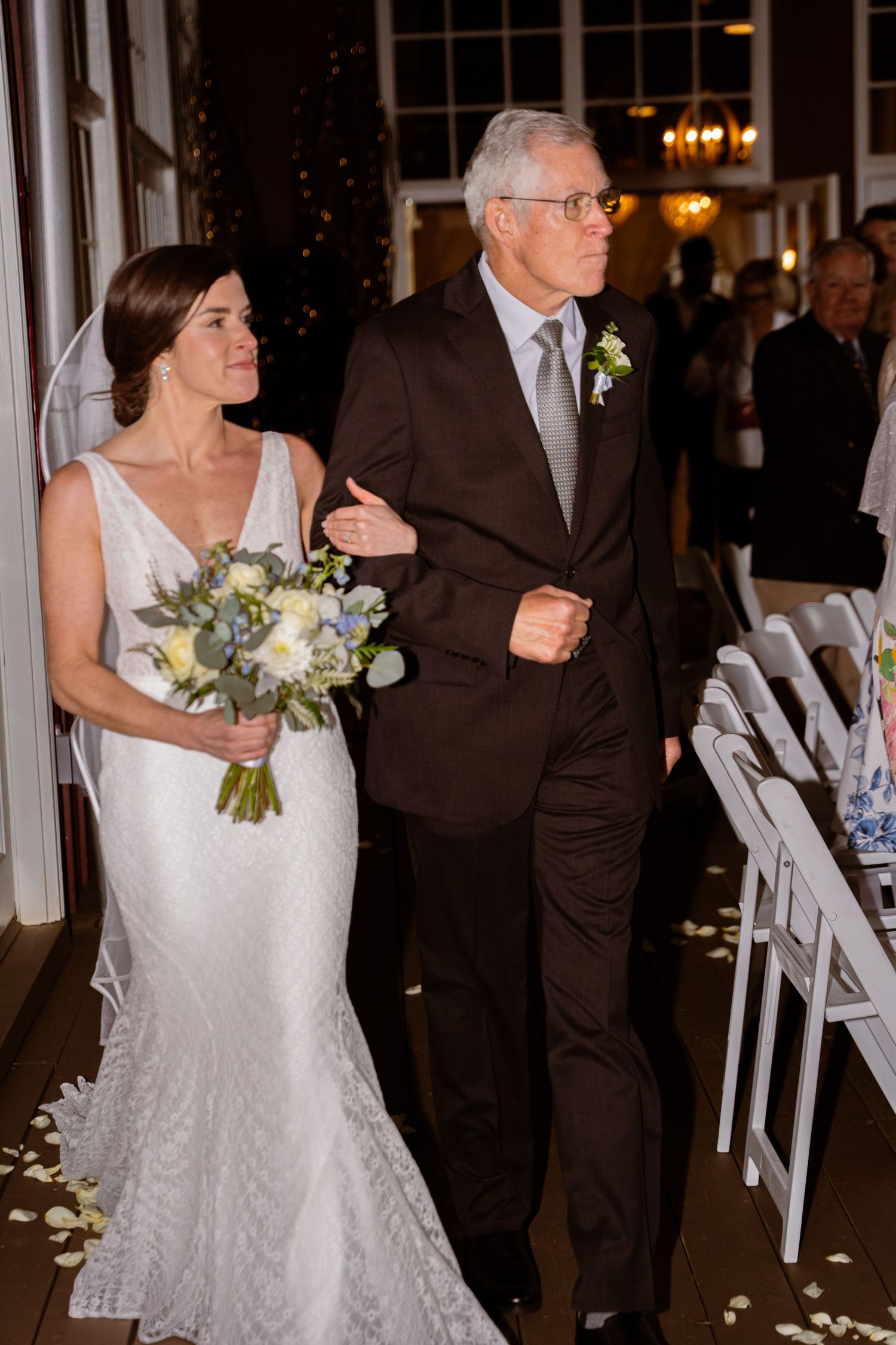 A bride in a lace wedding dress walks down the aisle with an older gentleman in a dark suit at an indoor ceremony.