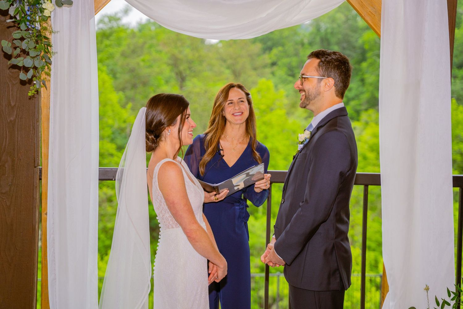 Wedding ceremony under white fabric gazebo with officiant in navy blue dress conducting service on outdoor deck.