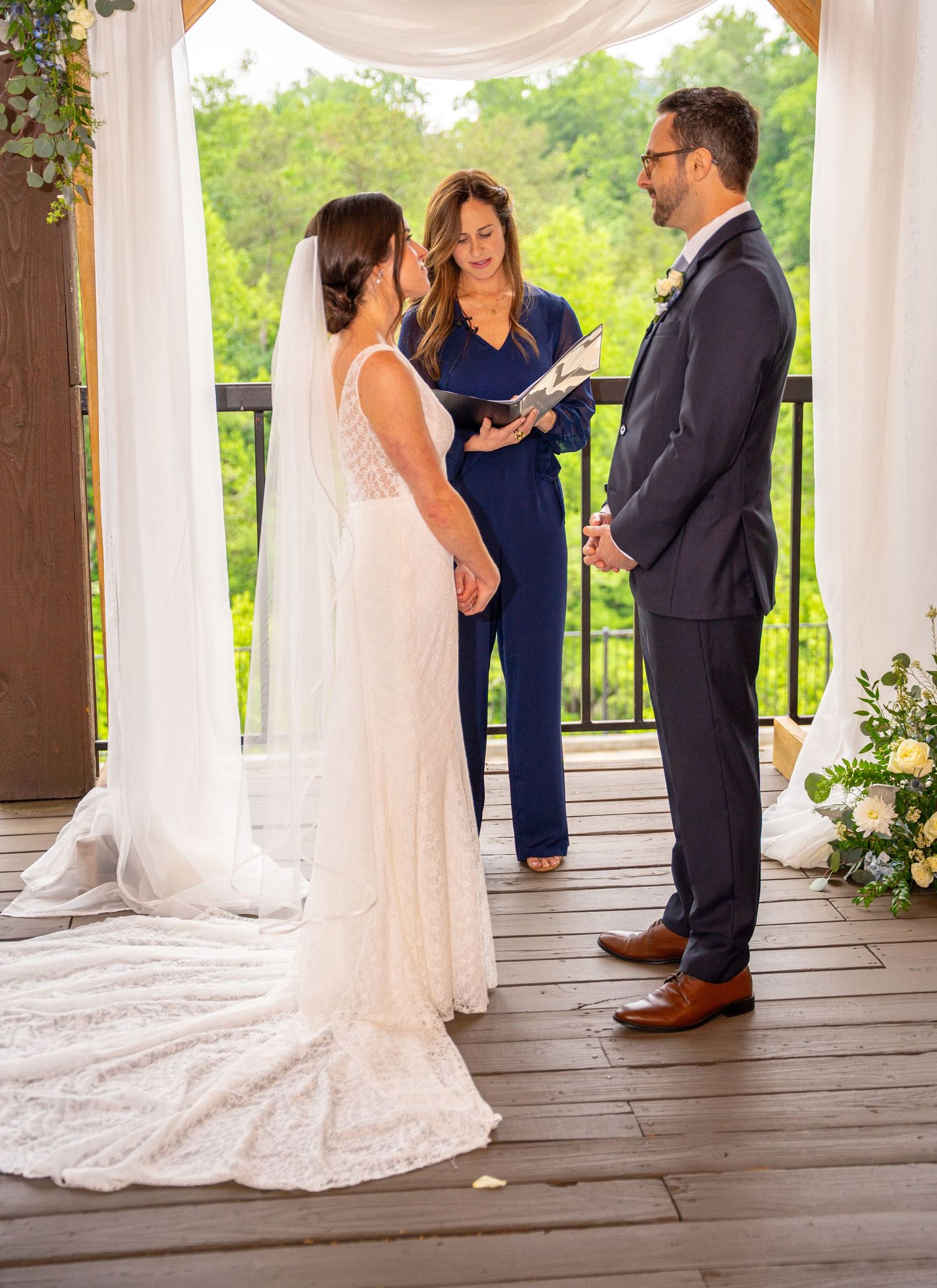 A wedding ceremony takes place on a covered deck with white draping and floral decorations in an outdoor setting.