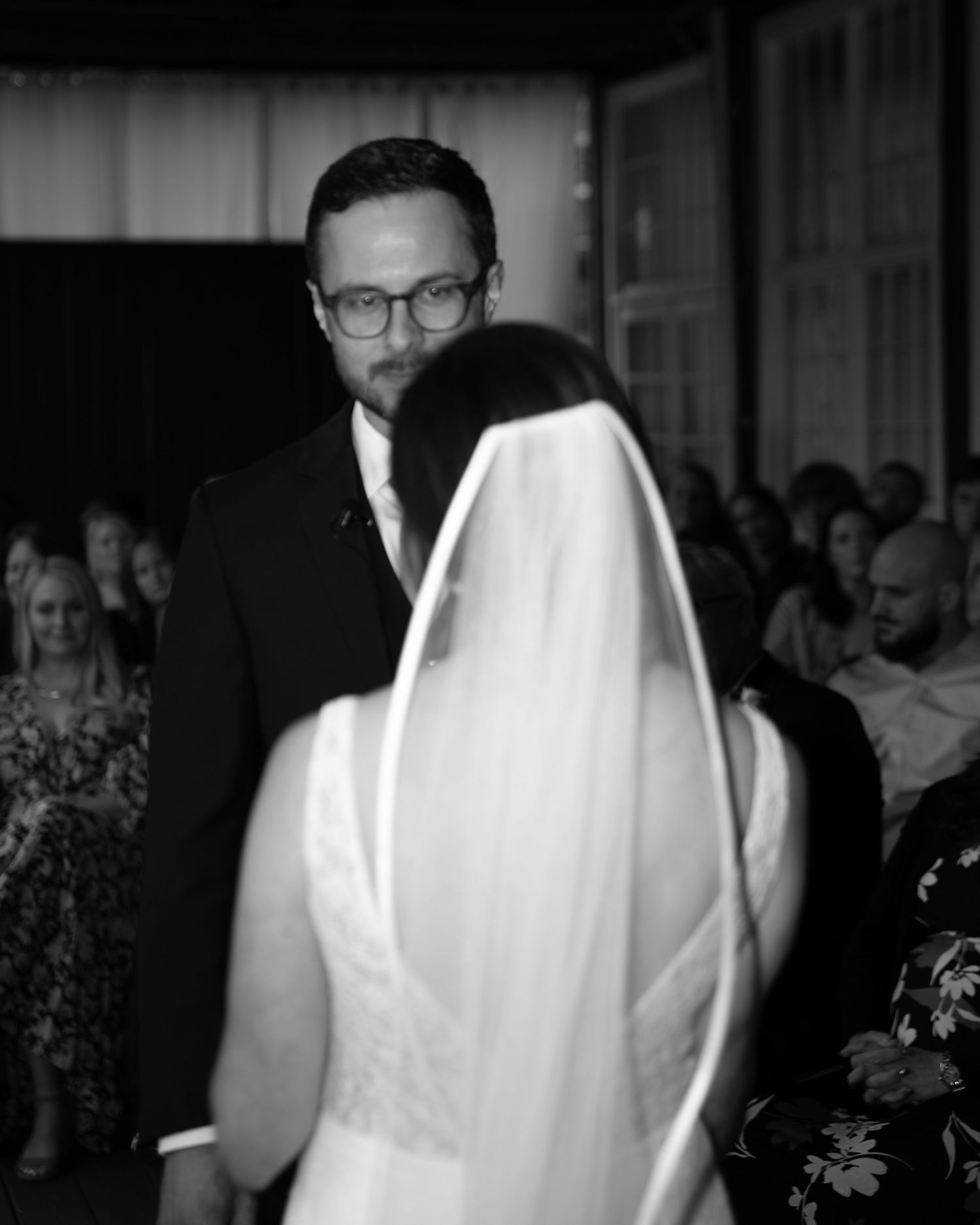 Black and white wedding ceremony photo showing joyful couple exchanging vows with guests in background.