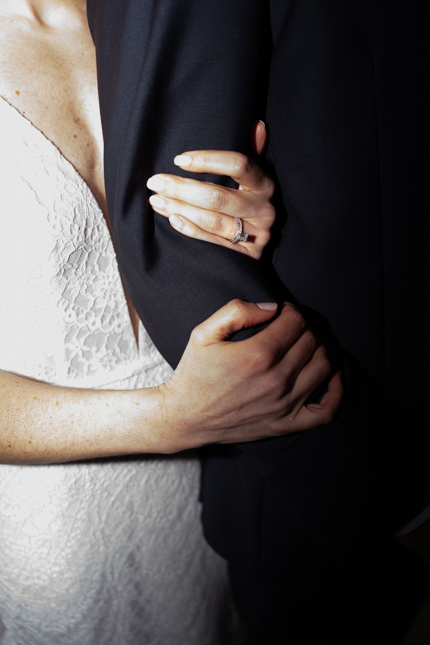 Close-up of a bride in white lace dress holding onto groom's black suit jacket.