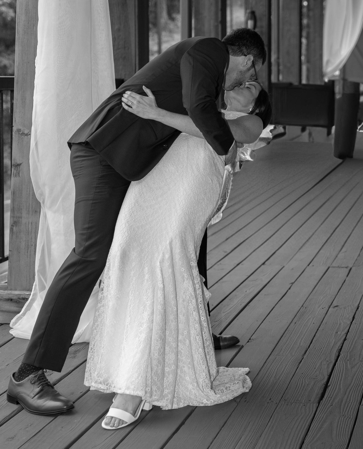 A romantic black and white wedding photo shows a couple embracing in a deep dip kiss on a wooden porch.