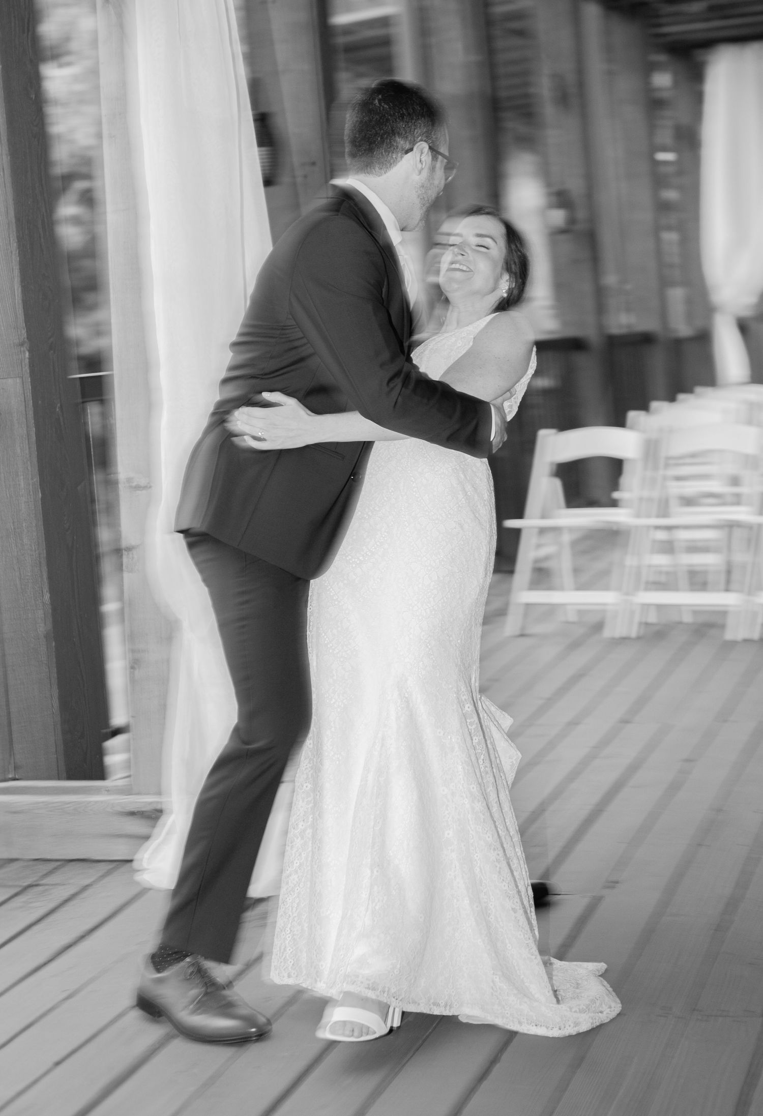 A black and white wedding photograph showing a couple sharing a joyful dance moment together on a porch.