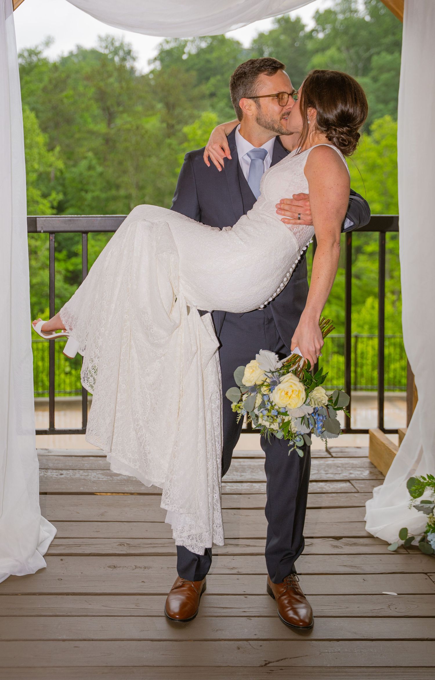 A couple in wedding attire share a romantic moment on a deck overlooking a lush forest landscape.