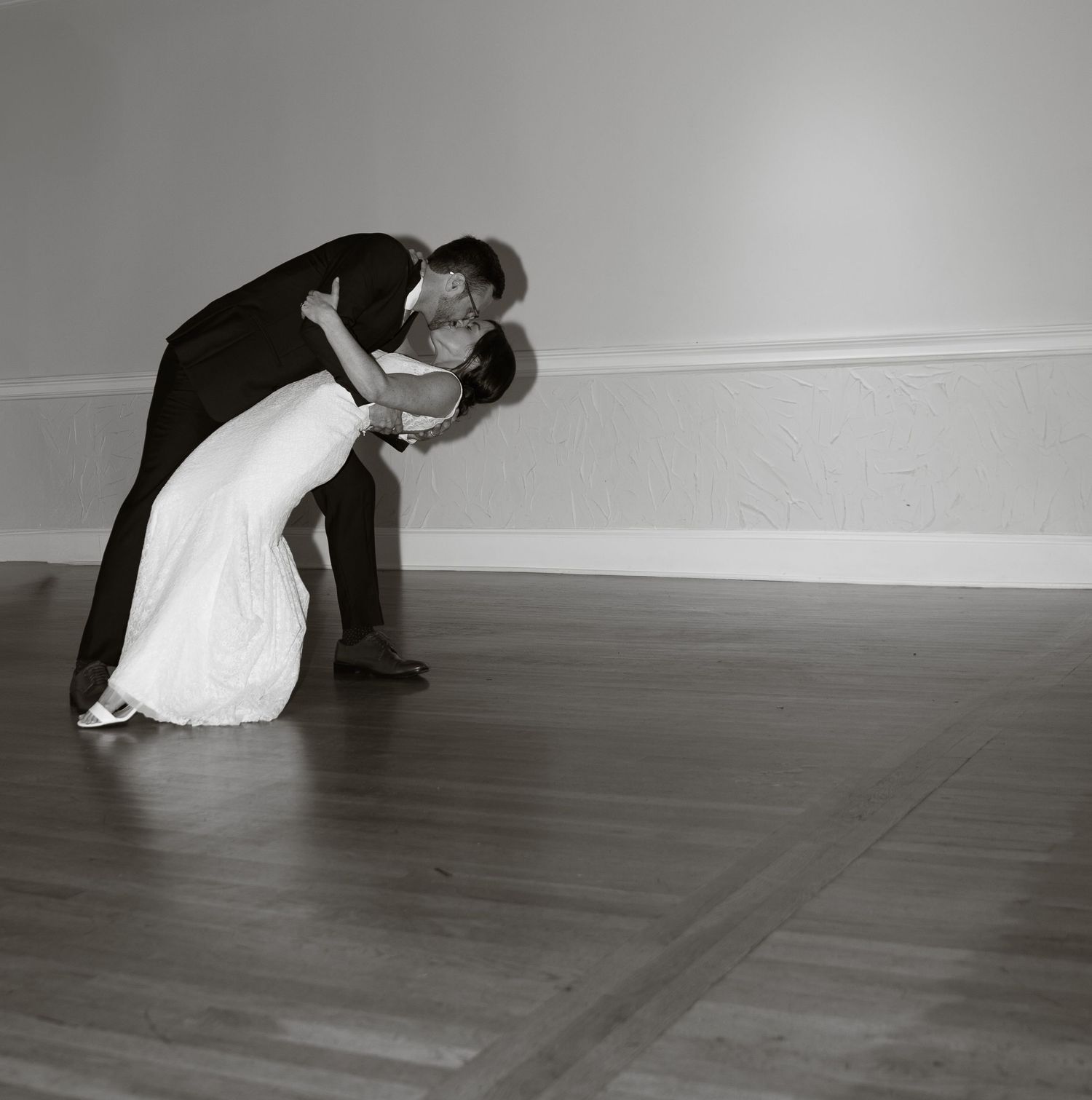 A couple in formal wedding attire performs a dramatic dance dip on a hardwood floor in black and white photography.