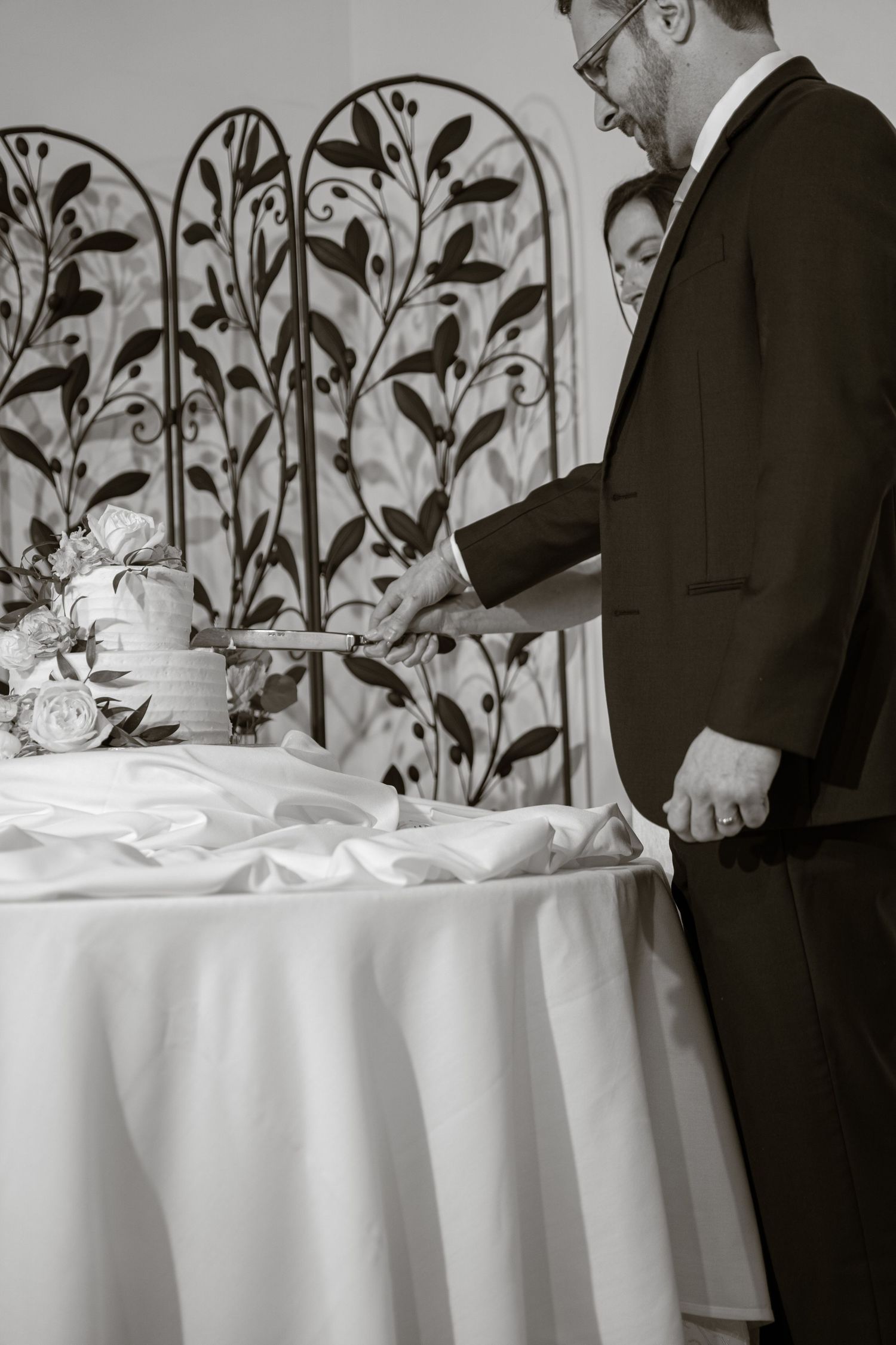 A black and white photo of someone cutting a wedding cake with decorative floral screens in the background.