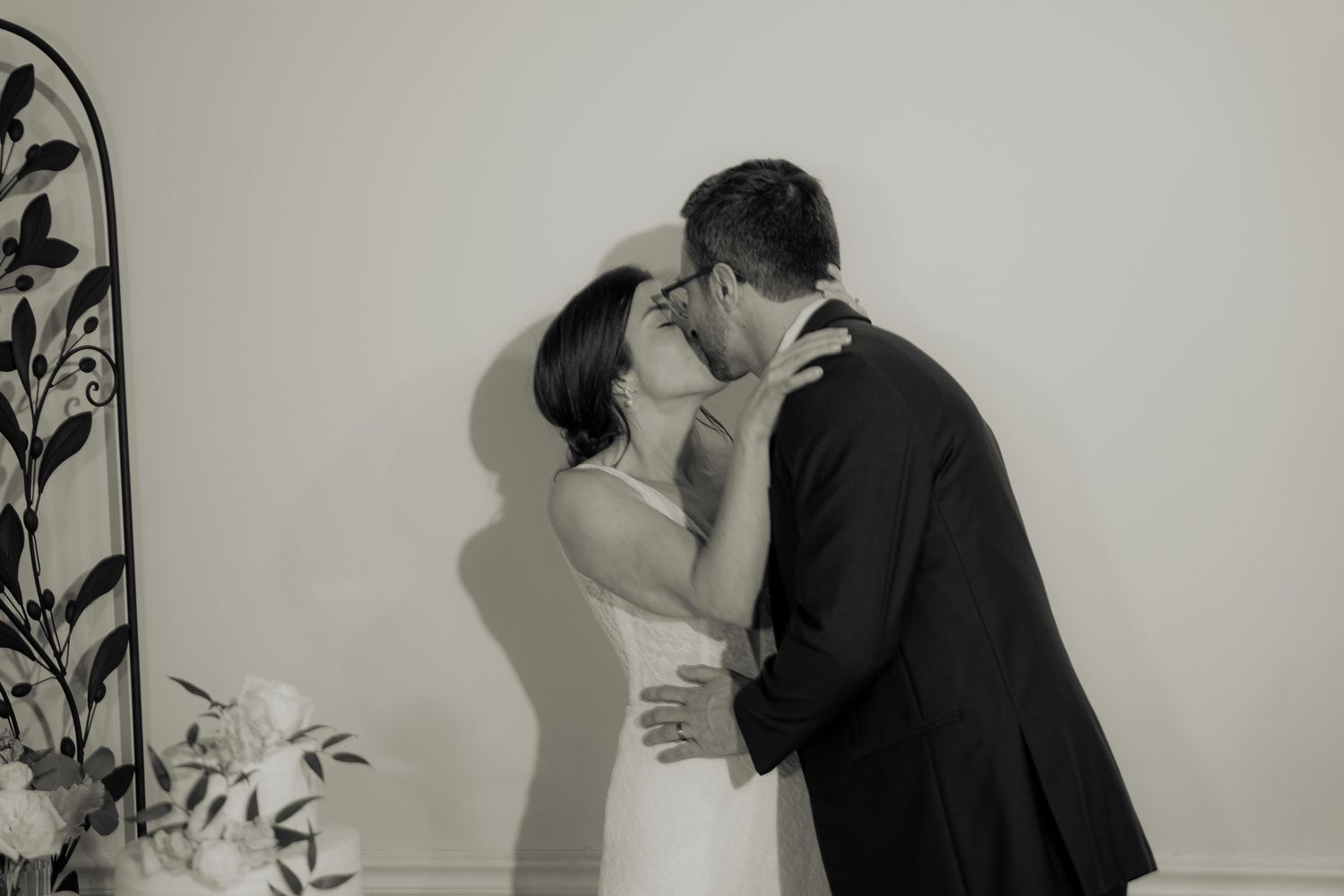 A romantic black and white wedding photo of a couple sharing a kiss against a plain wall with decorative foliage.