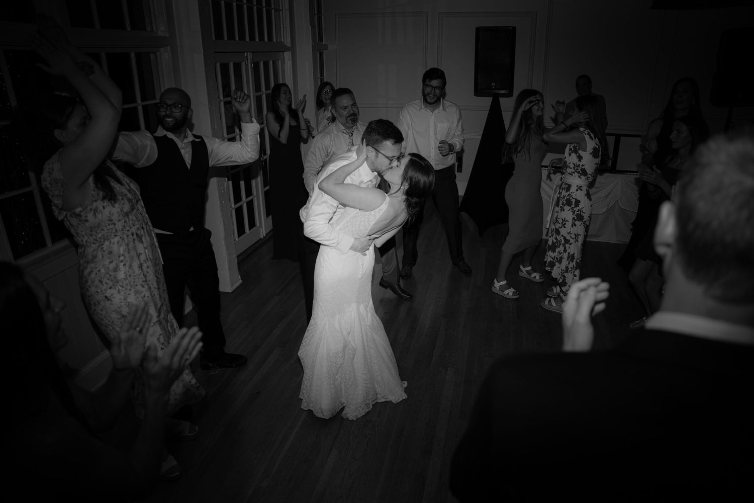 A couple shares a romantic slow dance in black and white at their wedding reception surrounded by guests on the dance floor.