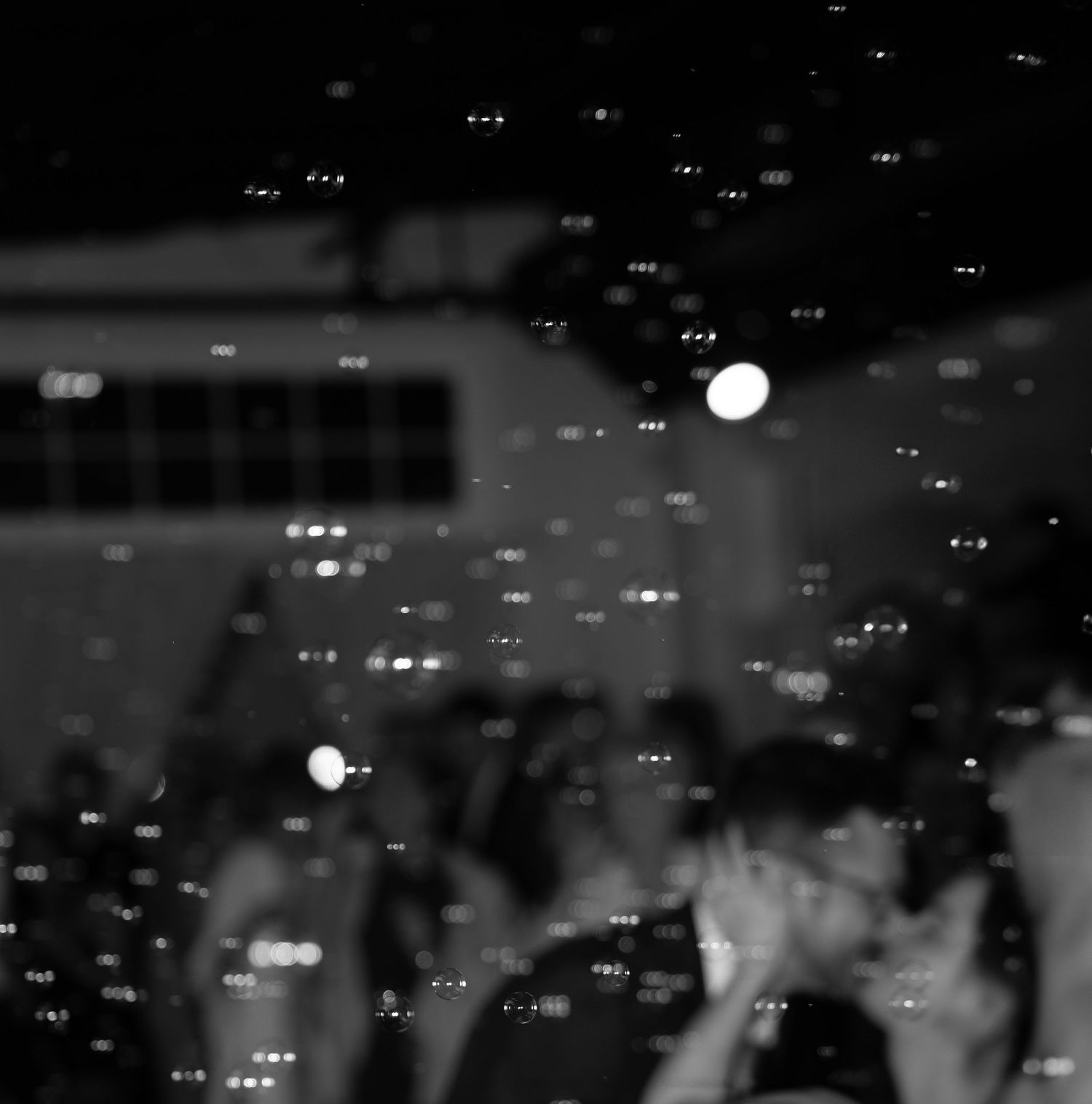 A black and white concert scene with bokeh lights and raised hands silhouetted against a dark background.
