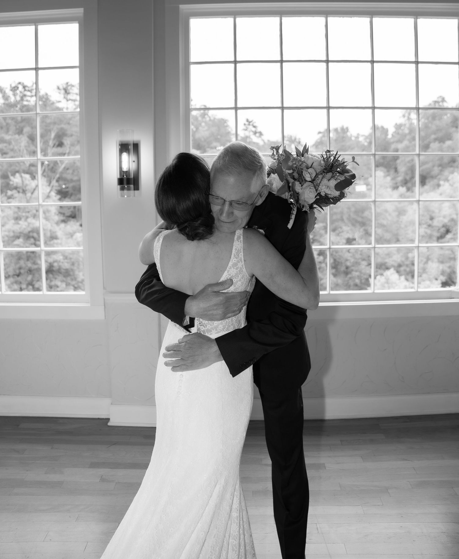 A couple embraces in a tender dance moment in front of large windows in a bright, elegant room.