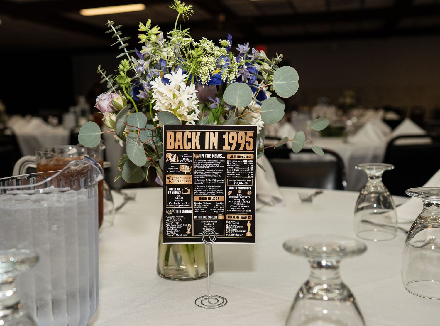 Wedding centerpiece featuring fresh flowers in a glass vase with a 'Back in 1995' sign on an elegant white tablecloth.
