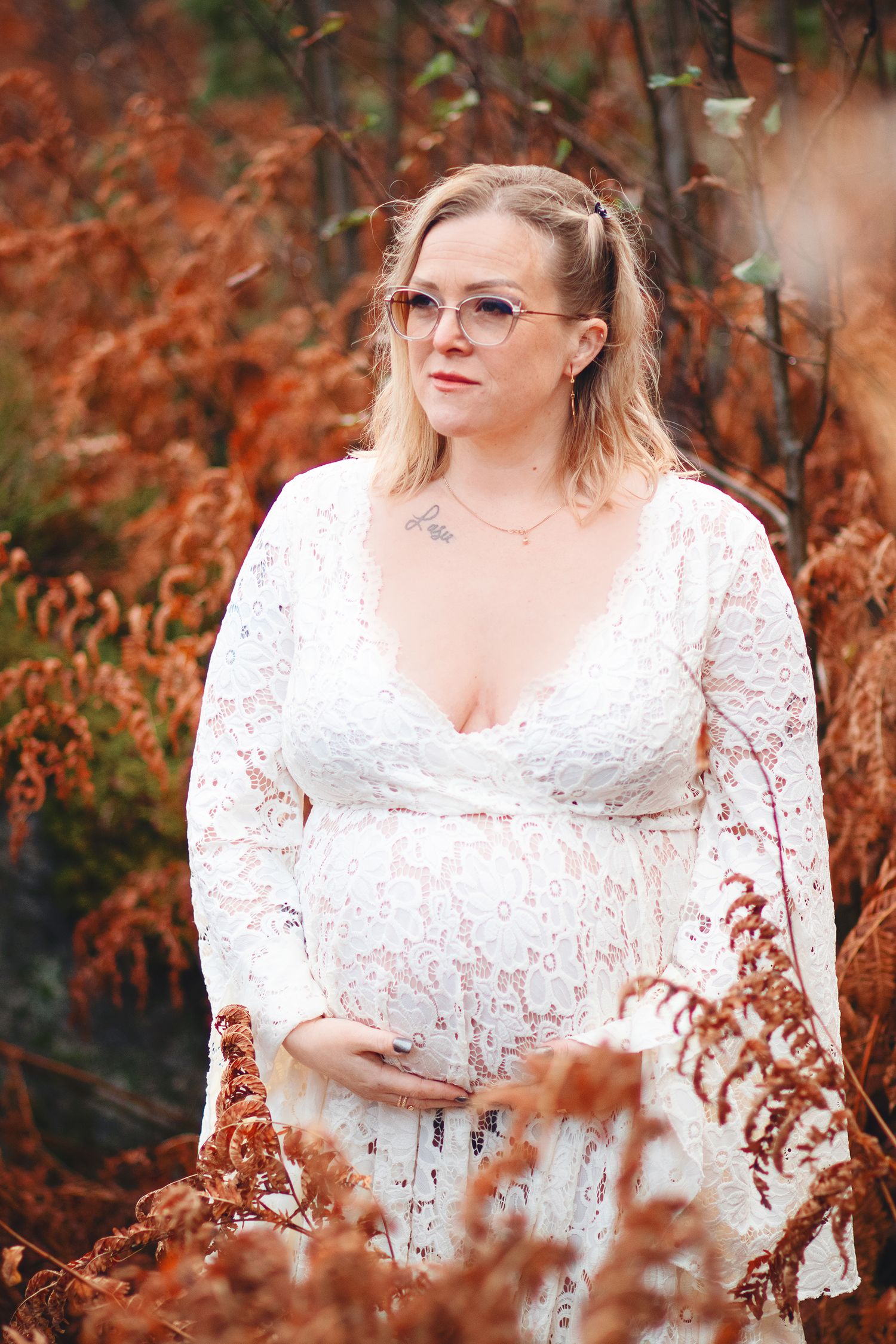 Pregnant woman in white lace dress with glasses standing among autumn red ferns outdoors.