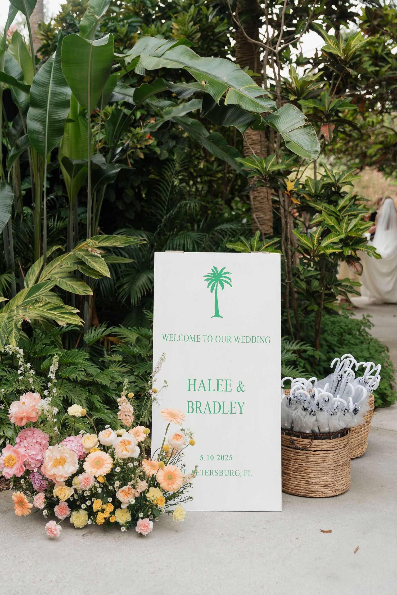 Elegant wedding welcome sign with palm tree logo amid tropical plants and peach-colored floral arrangements.