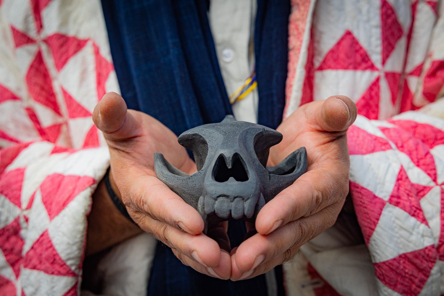 Hands gently hold a dark gray ceramic or 3D printed skull against a pink and white patterned fabric background.