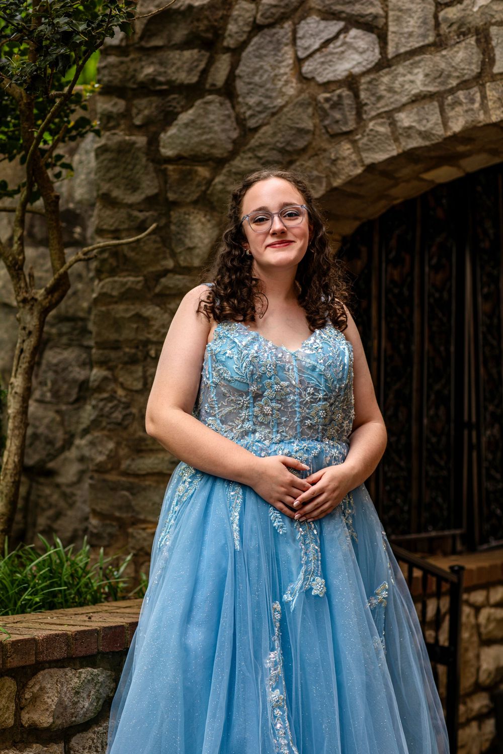 A person in a light blue formal gown with floral lace details poses in front of a rustic stone archway.