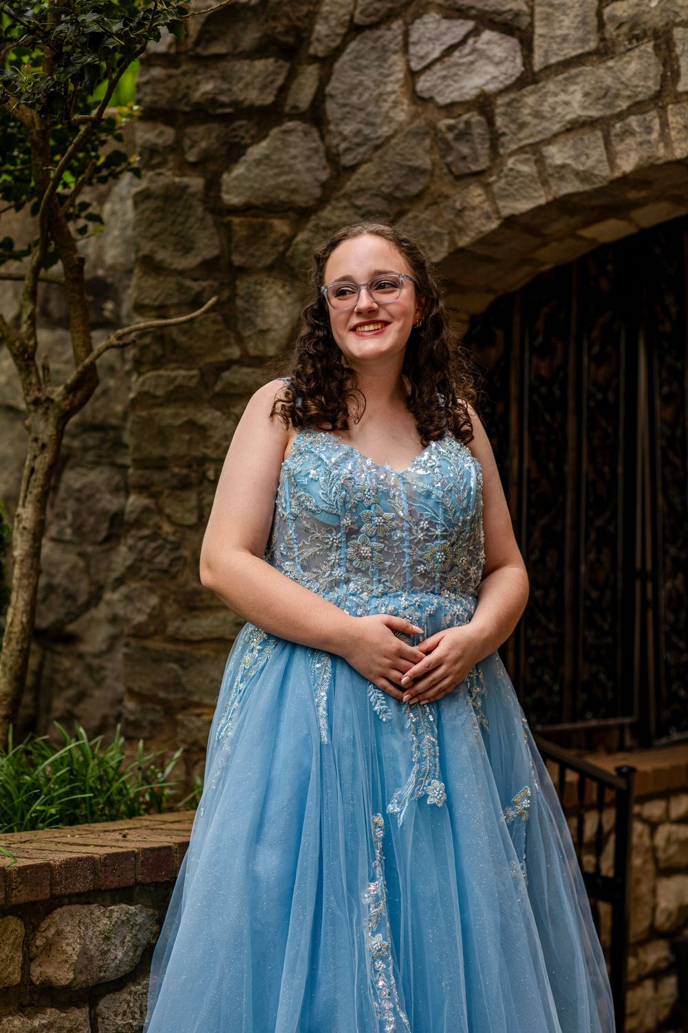 A person poses in a light blue formal gown with lace details in front of a stone archway.