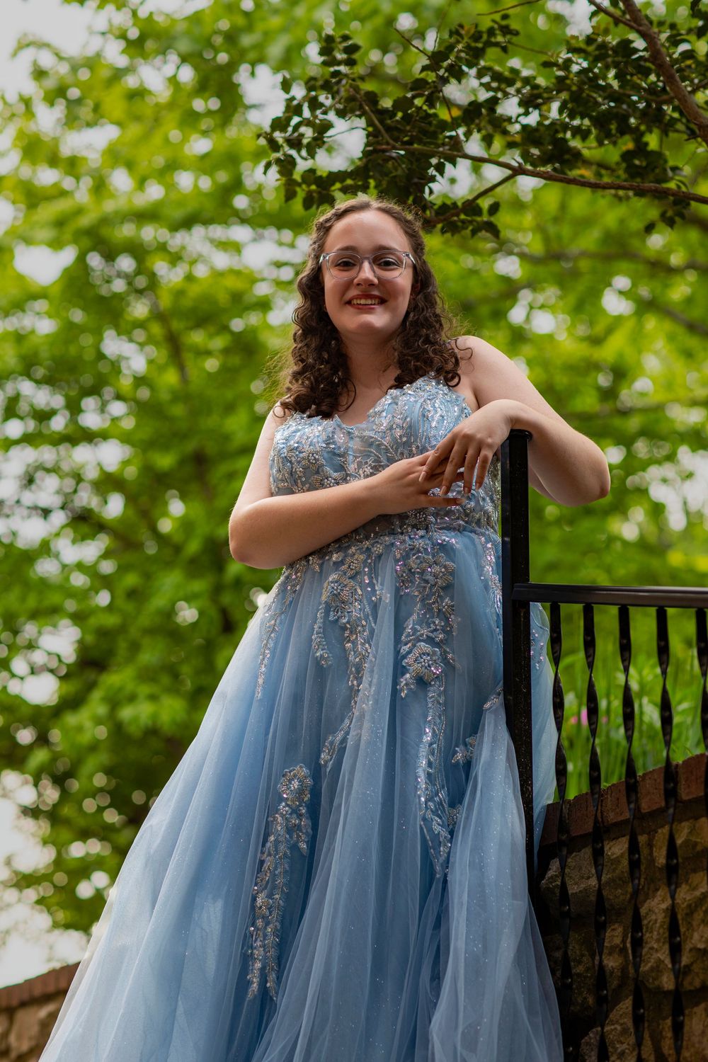 A person in a flowing blue and white embellished ballgown poses against lush green foliage.