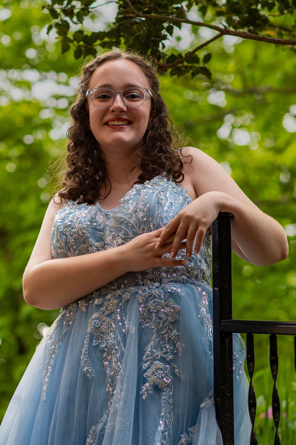 A person in a light blue formal gown with silver embellishments poses outdoors against a verdant background.