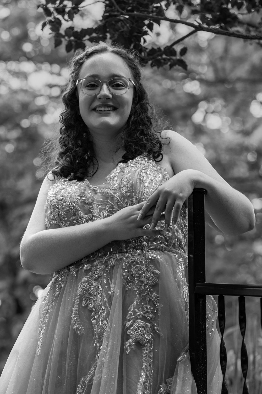 A black and white portrait of someone in a formal gown leaning against an outdoor railing.