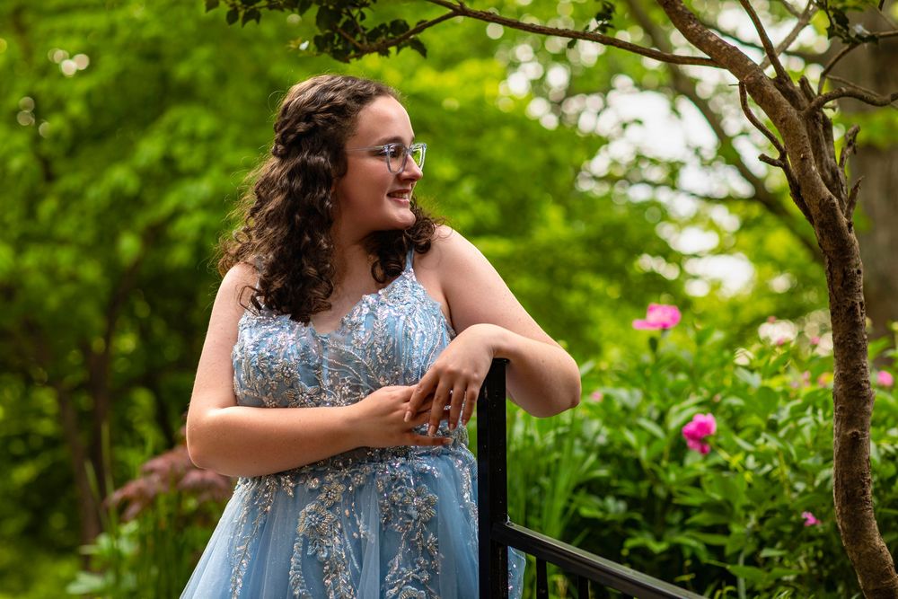 A person wearing a pale blue formal dress stands outdoors in a garden setting with green trees and pink flowers.
