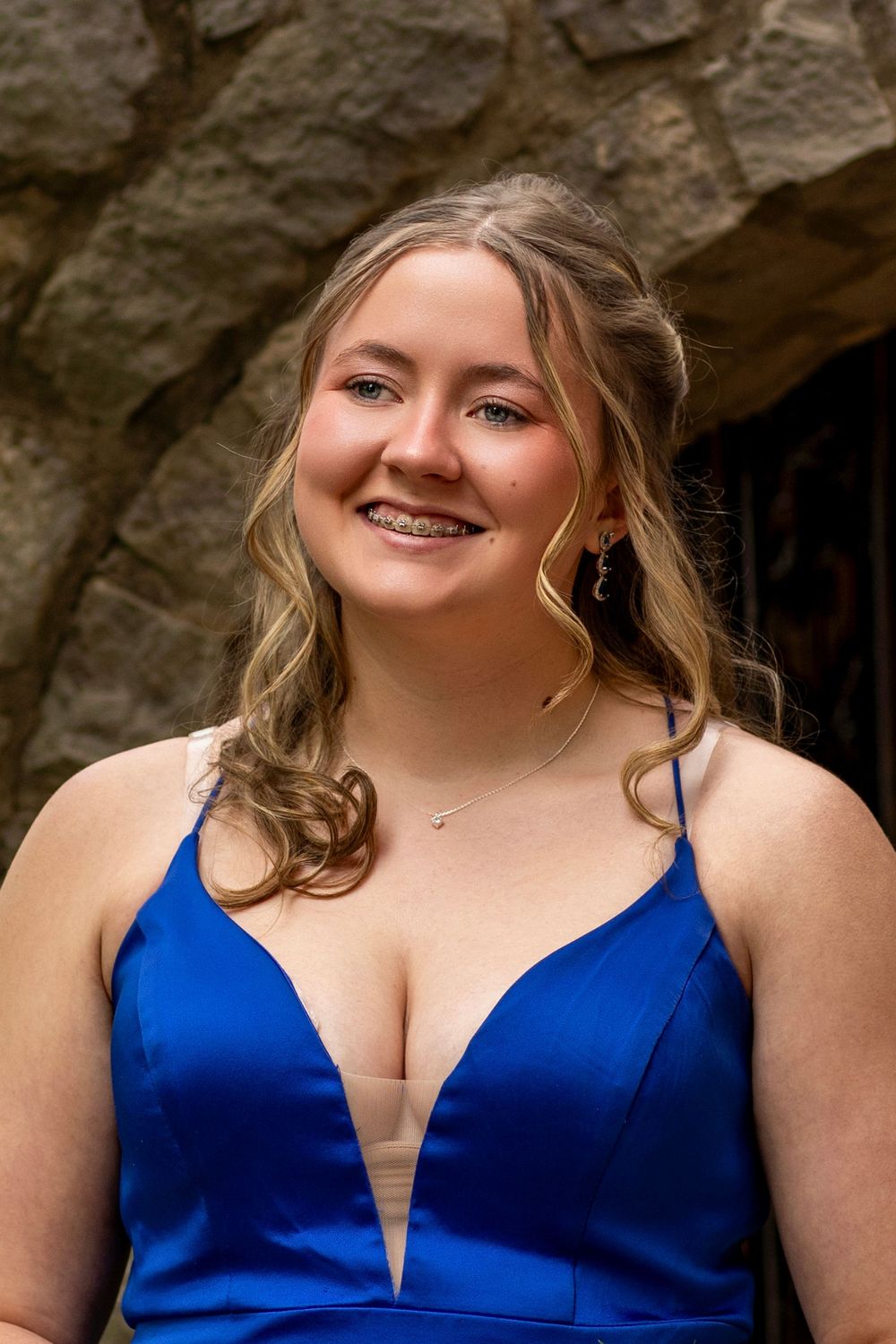A person in a royal blue formal dress with a plunging neckline smiles warmly against a stone backdrop.