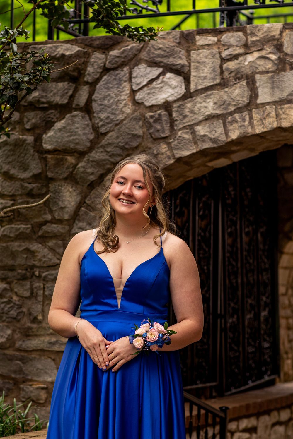 A woman in an elegant royal blue formal gown poses by a stone archway with a corsage.