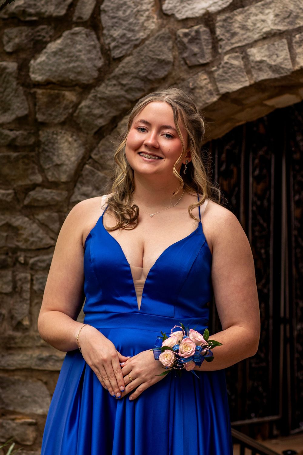 A person in a vibrant royal blue formal dress poses outdoors against a stone wall with a peach and blue floral corsage.