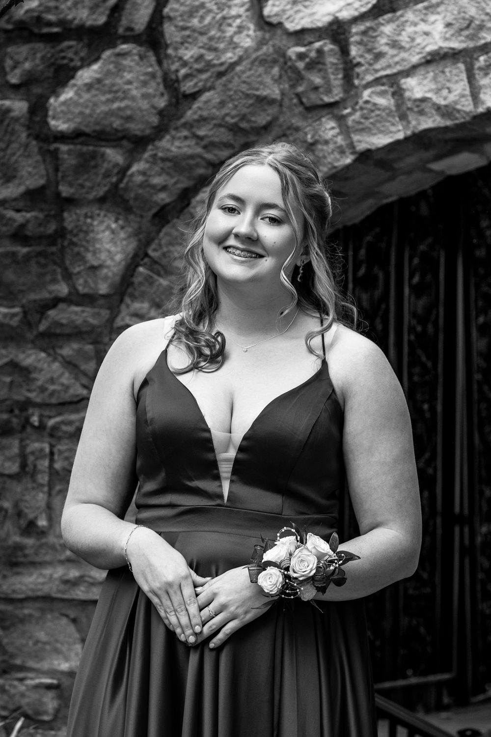 A woman in a black formal dress holds a corsage while standing in front of a stone wall during a special occasion.