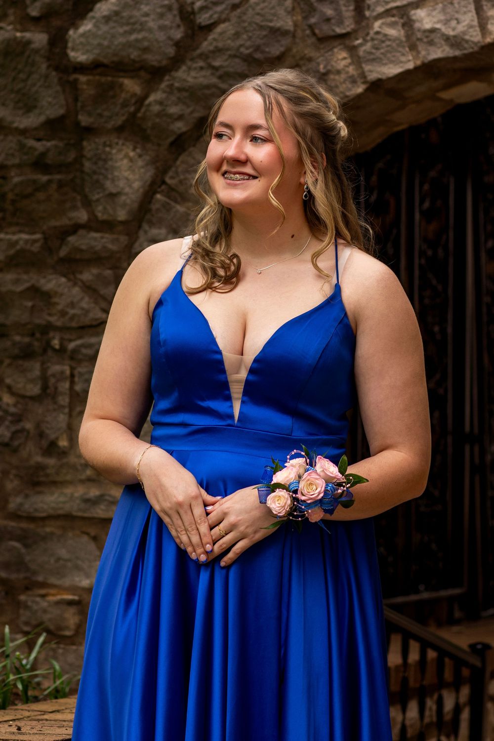 A person in a royal blue formal dress poses in front of a stone wall with floral wrist corsage and jewelry.