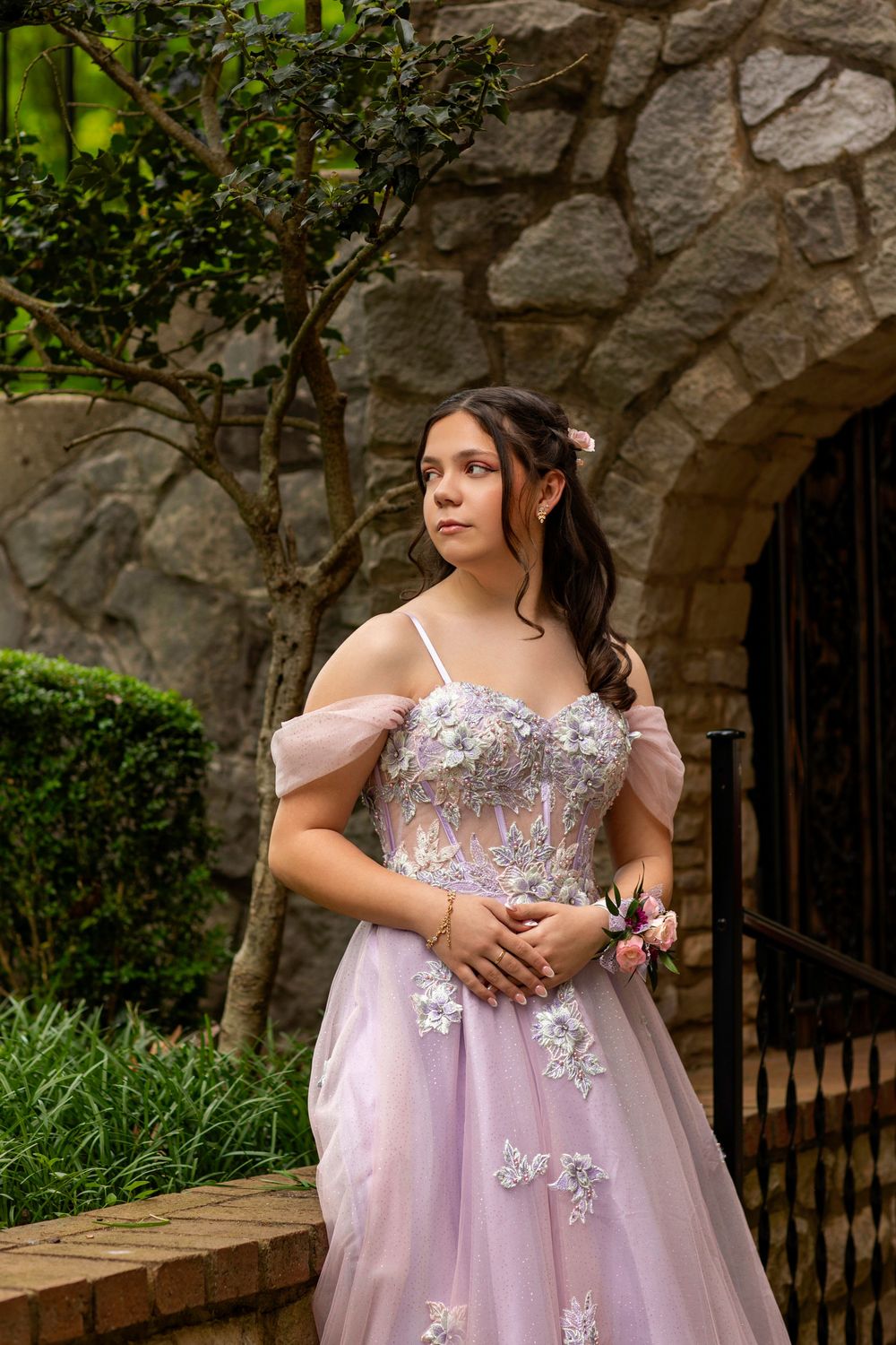 Person wearing a lavender tulle prom dress with floral lace appliques poses by stone archway and garden.