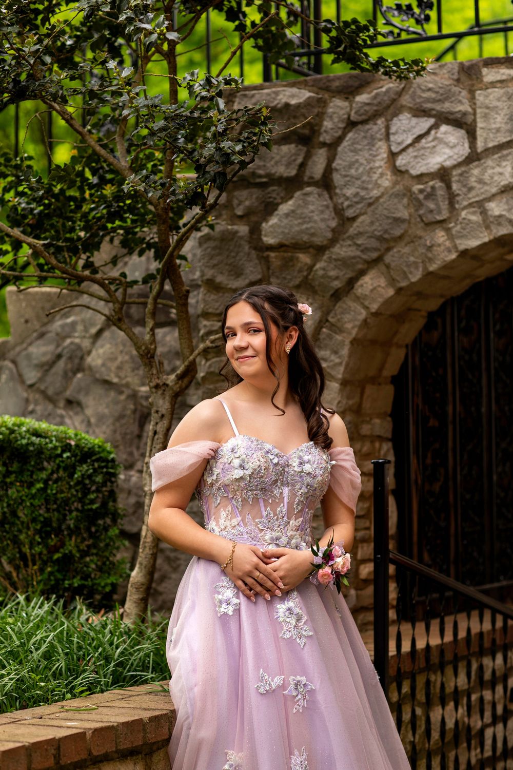 Off-shoulder pink tulle gown with floral appliques photographed in front of stone archway with decorative iron gate.