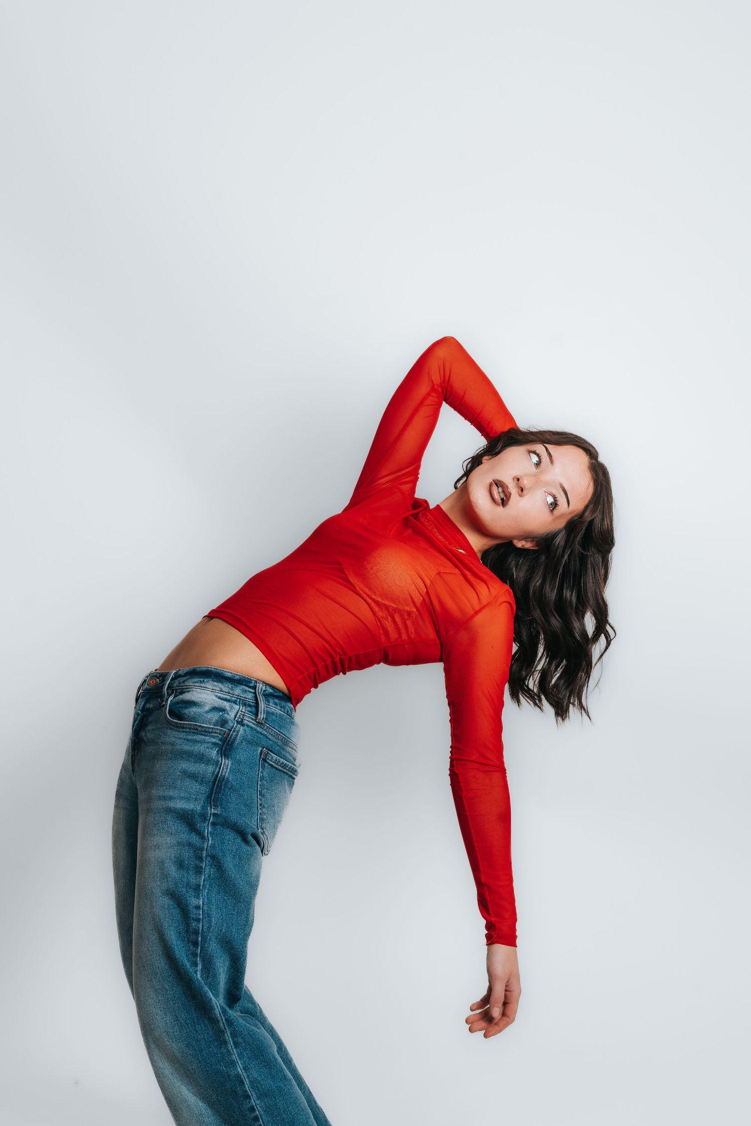 Model in red long sleeve top and jeans poses against white wall in dynamic arching pose.