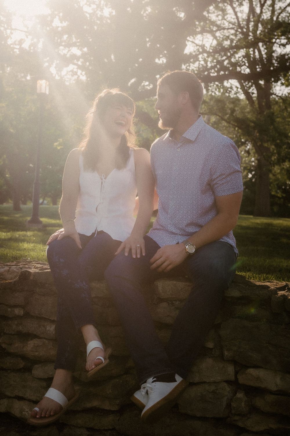 A couple sits on a stone wall at sunset in a park setting with sun rays filtering through trees behind them. Loose Park Kansas City.