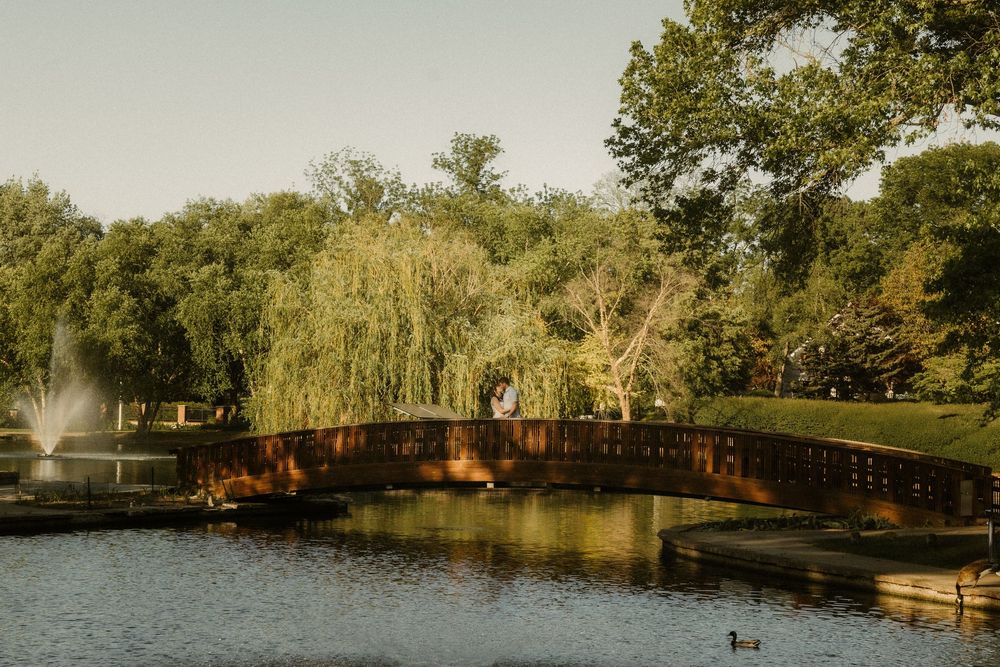 A scenic park bridge spans over a pond with a fountain and willow trees in the background with couple kissing on bridge.