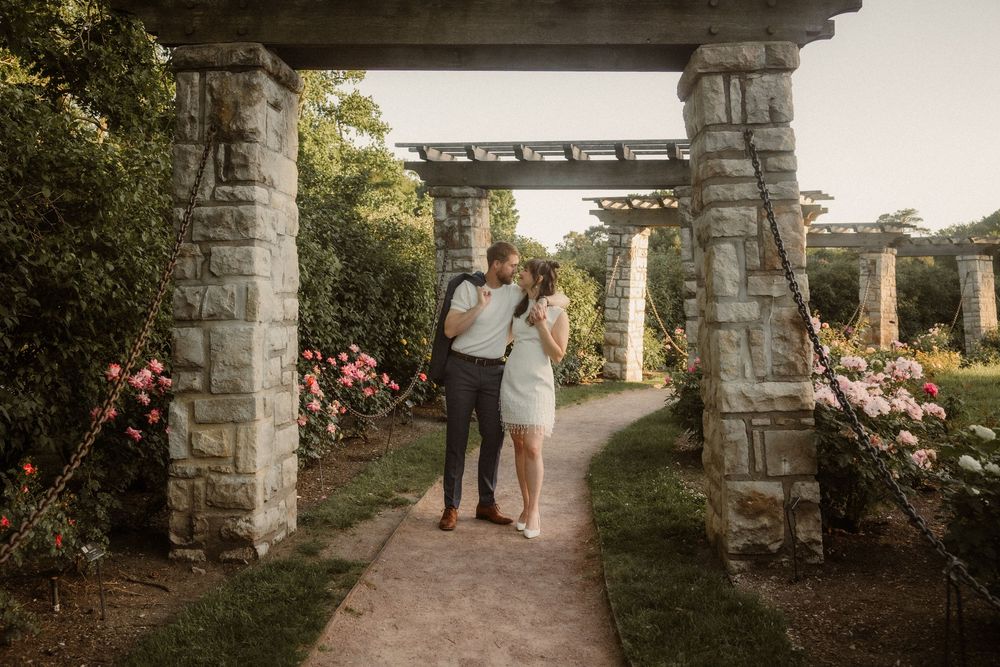 Two people stand together on a garden path beneath stone archways surrounded by blooming flowers at sunset. Engagement Photo at Rose Garden.